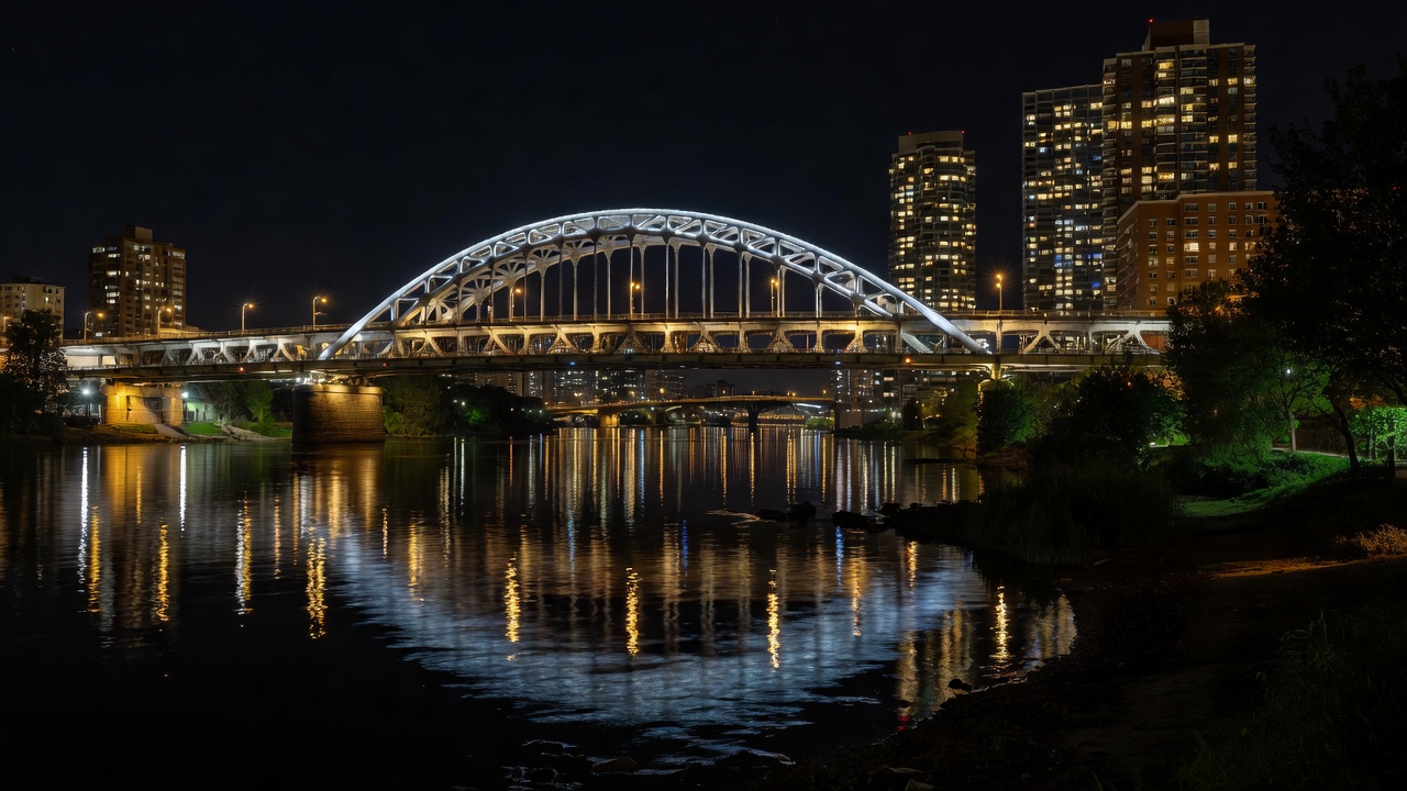 An illuminated urban bridge viewed with nearby buildings, dark water and a quieter riverbank